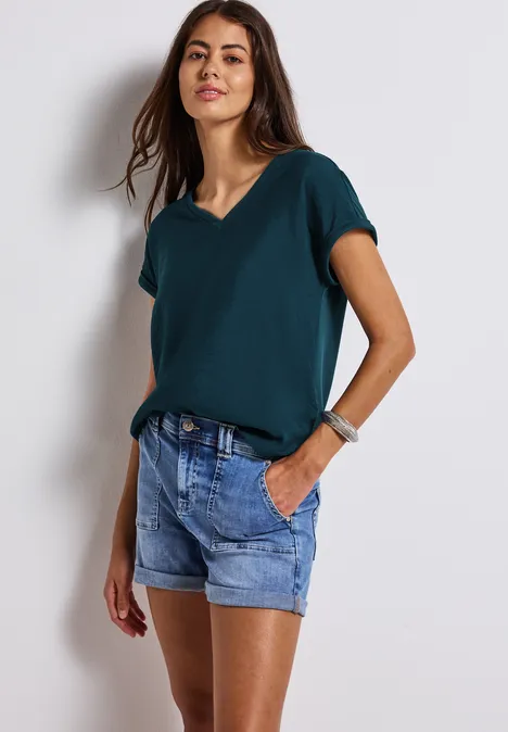 Frau mit dunkelgrünem T-Shirt und Jeansshorts steht vor weißer Wand.